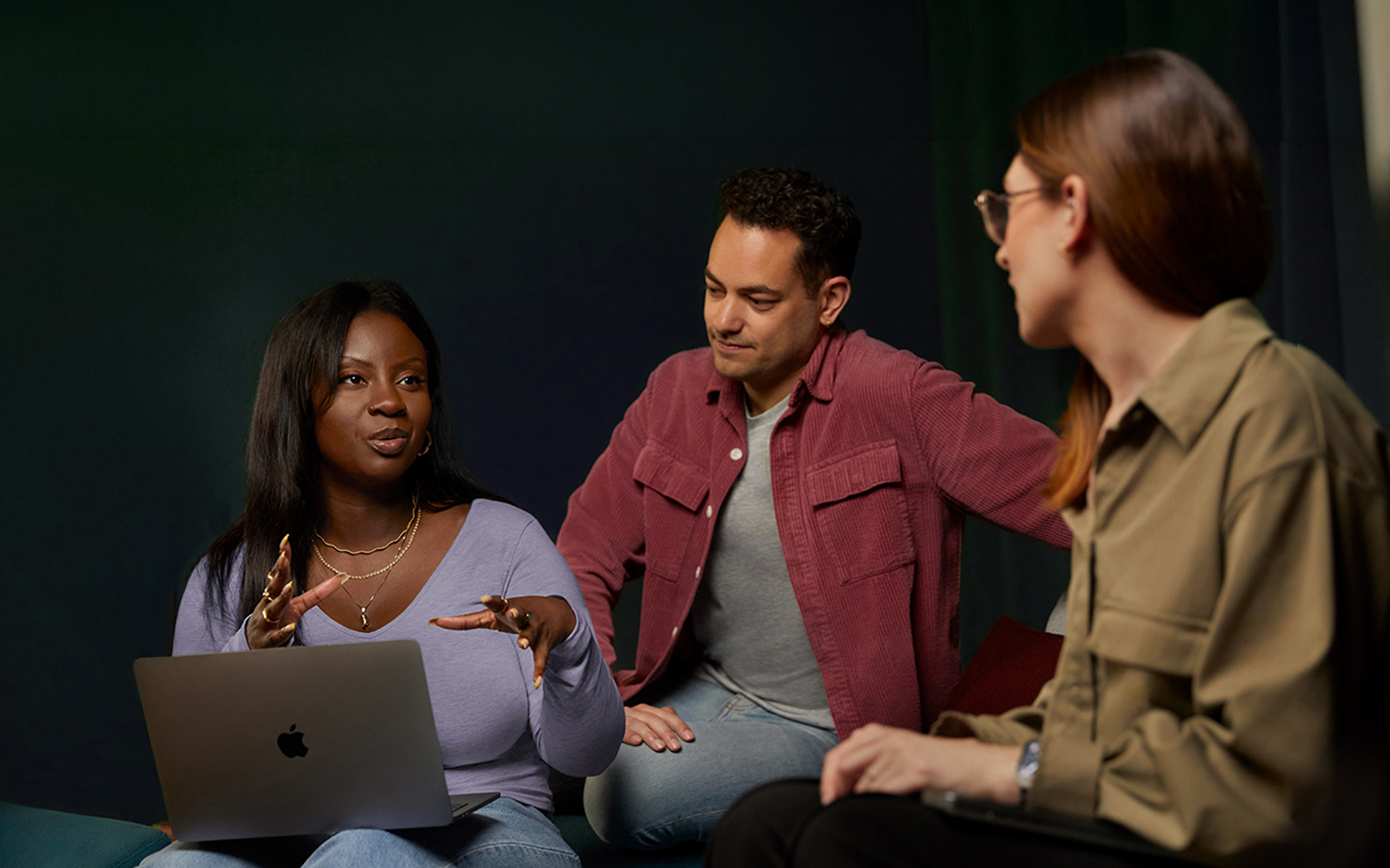 Tres personas del equipo de Software y servicios de Apple conversan en un sillón, una de ellas con una MacBook.