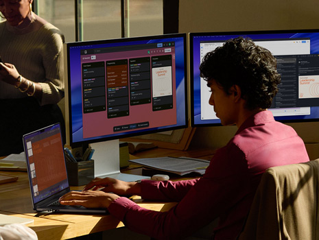 Equipo de empleados sentados alrededor de una mesa en un entorno de oficina. Una mujer sentada en su escritorio trabaja en un MacBook y varios monitores. Sus colegas están reunidos charlando.