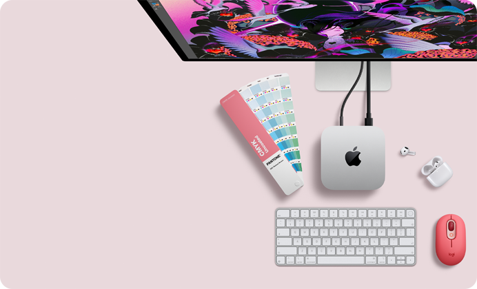 Skats no augšas uz Mac mini sudraba krāsas alumīnija korpusā ar noapaļotiem stūriem un spīdīgu melnu Apple logotipu centrā, savienots ar vadu. Augšpusē daļēji redzams Studio Display, atvērti AirPod 4, Pantone krāsu paletes, cipartastatūra un pele