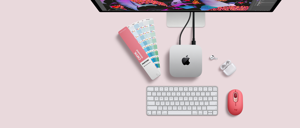 Pohľad zhora na strieborný hliníkový Mac mini so zaoblenými rohmi a lesklým čiernym logom Apple uprostred. Káblom je pripojený k displeju Studio Display, ktorý je čiastočne vidieť navrchu. Vedľa sú otvorené slúchadlá AirPods 4, vzorky farieb Pantone, numerická klávesnica a myš.