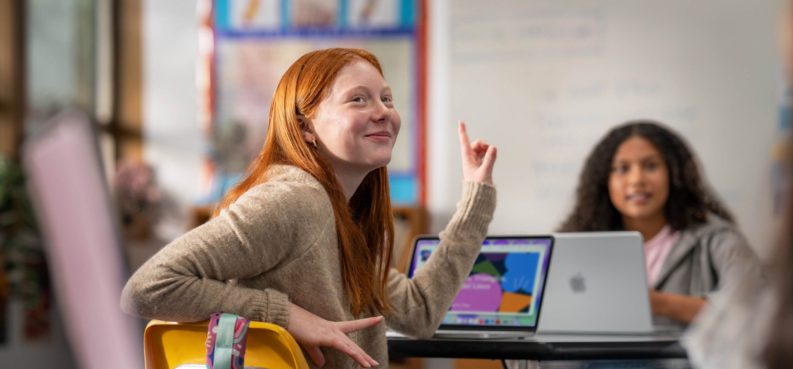Una estudiante sentada frente a una MacBook en un salón de clases levanta la mano para responder una pregunta, se muestra la facilidad de uso y la participación en clase