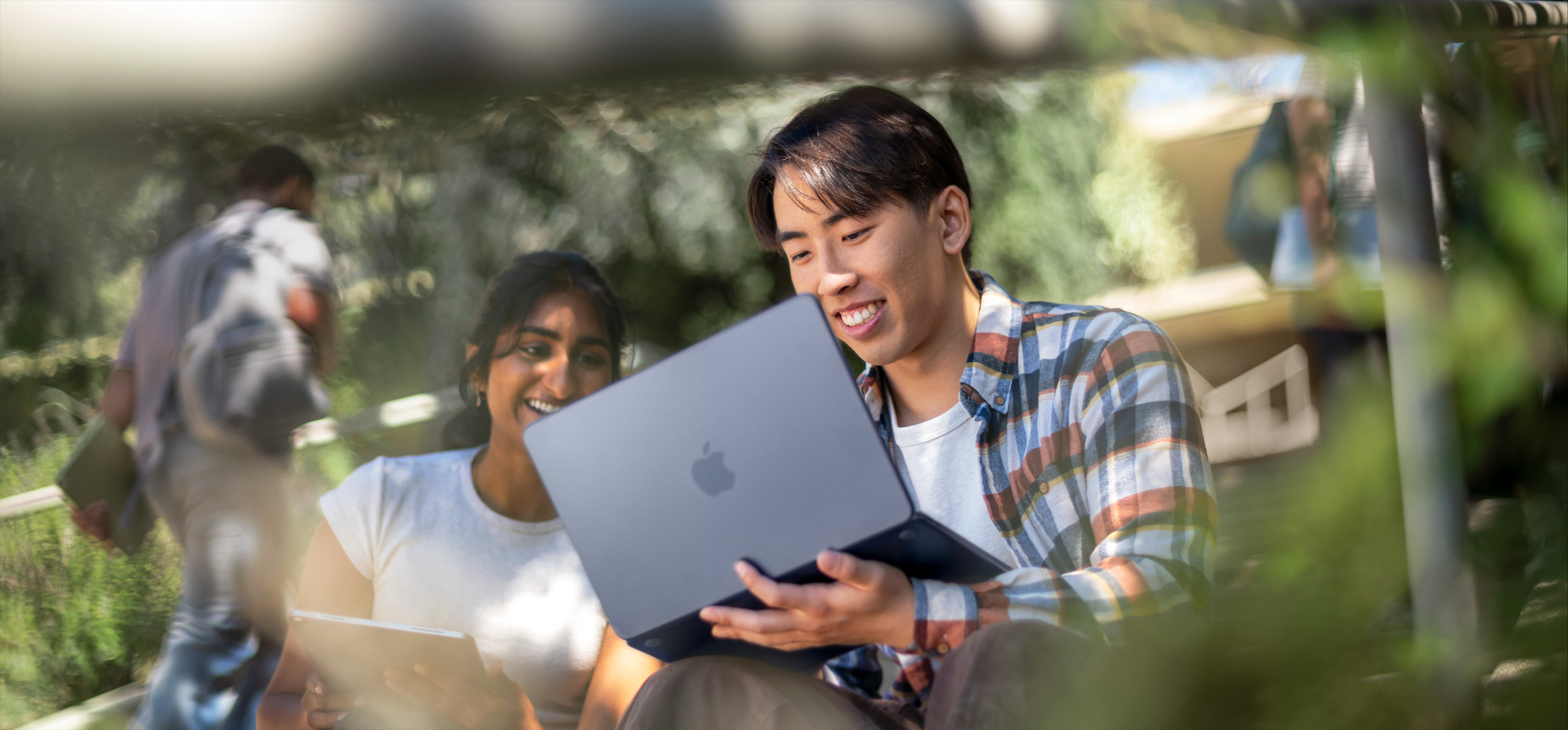 Dos estudiantes sonríen mientras usan una MacBook al aire libre, uno la sostiene en su regazo mientras colaboran en un proyecto