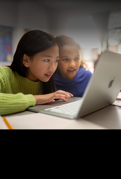Dos estudiantes de primaria en un escritorio miran con atención una MacBook mientras trabajan en un salón de clases