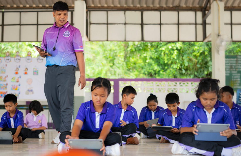 In an outdoor classroom, a physical education teacher walks among his students, who are seated on the ground using iPads.