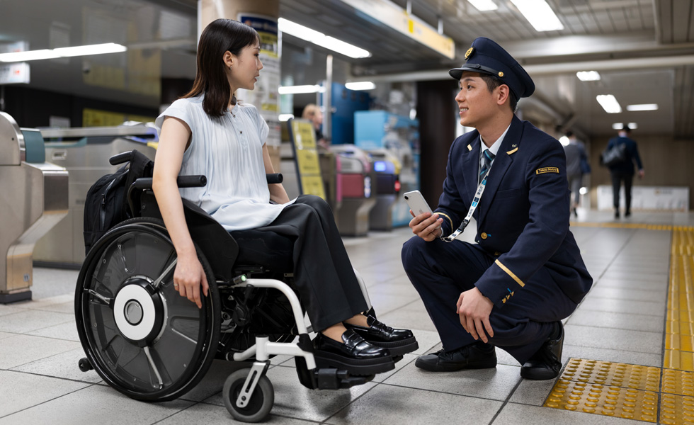 A professionally-dressed woman in a wheelchair and a station attendant have an exchange on the platform.