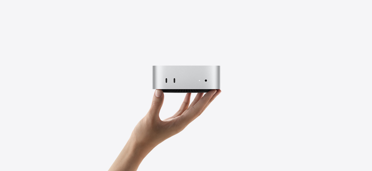 „Mac mini“ vaizdas iš priekio, laikomas ištiestos į viršų rankos pirštų galiukais, priekyje matomi du „Thunderbolt“ prievadai, būsenos indikatoriaus lemputė ir ausinių jungtis, juodos spalvos, apačioje siaurėjantis pagrindas, plokščias viršus, užapvalinti šonai, tiesūs kraštai, sidabro spalvos